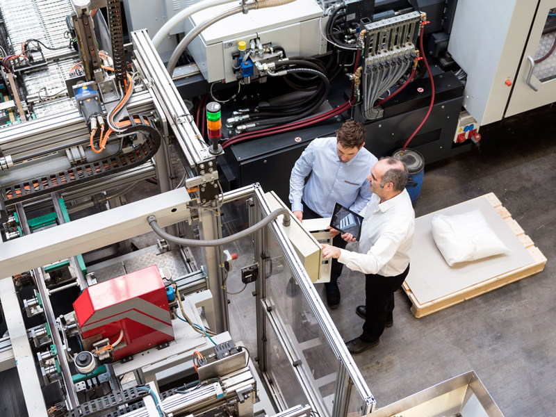 Two men discuss quality assurance and compliance requirements whilst looking at plastic manufacturing machinery